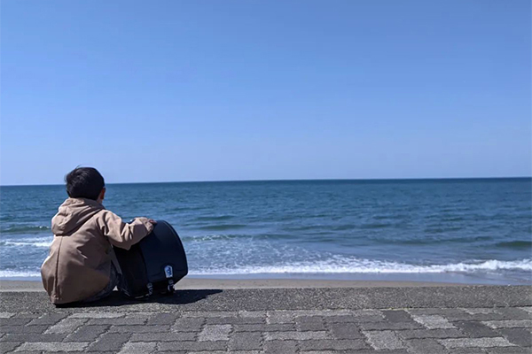 海を背景にランドセルと一緒に座ってる子どもの後ろ姿